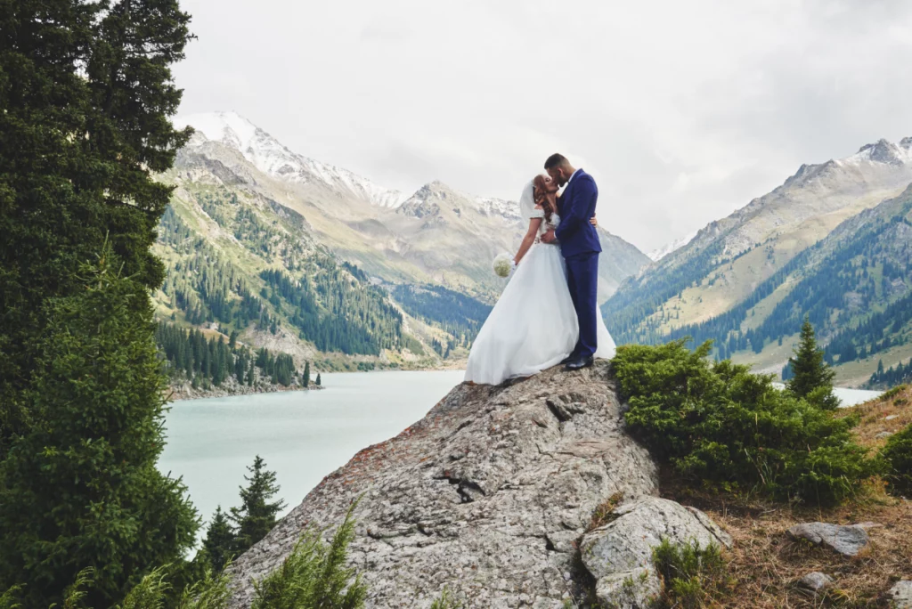 A newly web couple kissing in an embrace at the edge of a rocky cliff with a blue lake and majestic mountains in the background.