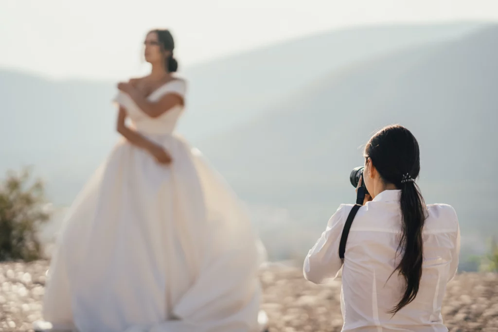 Photographer in the foreground with her back toward the camera in the forground while she photographs a soft focused bridge in her wedding dress with a mountain backdrop