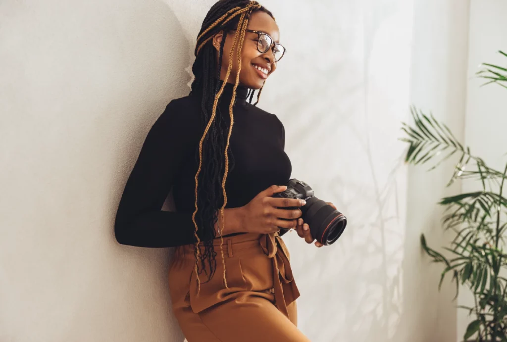 A photographer with long black and brown braids, wearing a black long-sleeve turtleneck and brown tie-waist pants, leans against a wall, smiling.