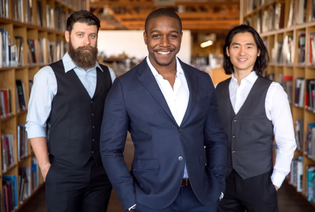 Three men stand in a library, wearing black, navy, and gray suits with white dress shirts.