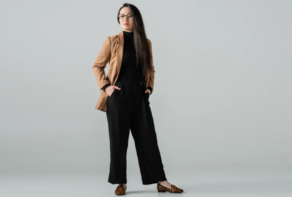 A woman with long brown hair stands in front of a gray background, wearing half-rim glasses, a black turtleneck, black wide-leg pants, a brown jacket, and leopard-print flats.