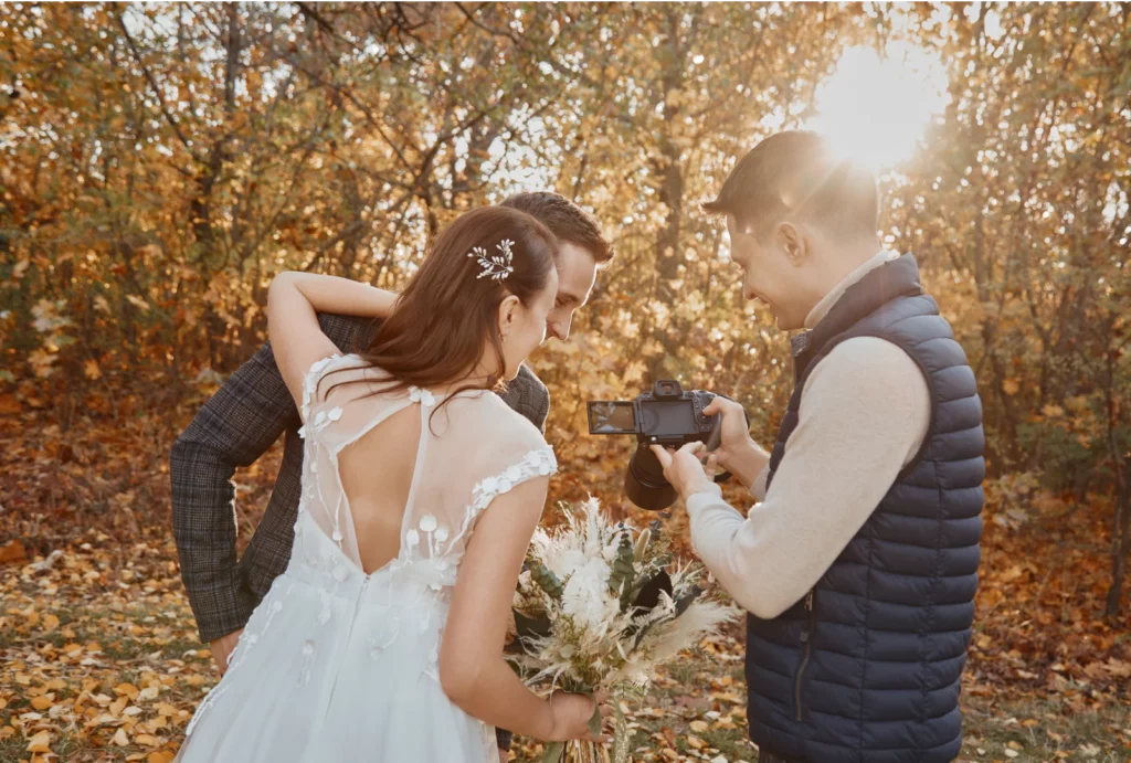 A photographer in a cream sweater and dark puffer vest reviews photos with a bride and groom during an outdoor photoshoot.