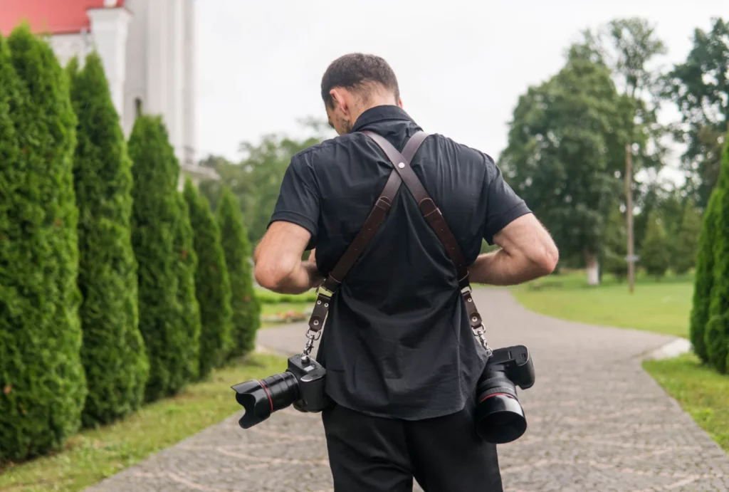 Back view of a professional photographer dressed in a black short-sleeve collared shirt and black slacks, carrying two cameras.