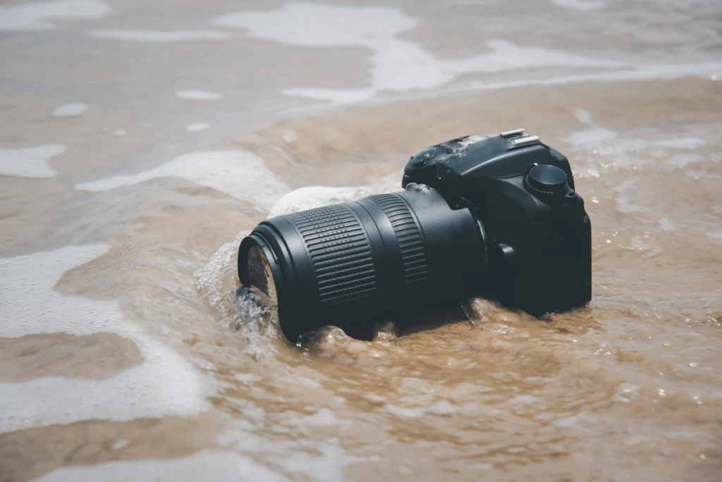 Close-up of a small wave hitting a black DSLR camera on a sand beach