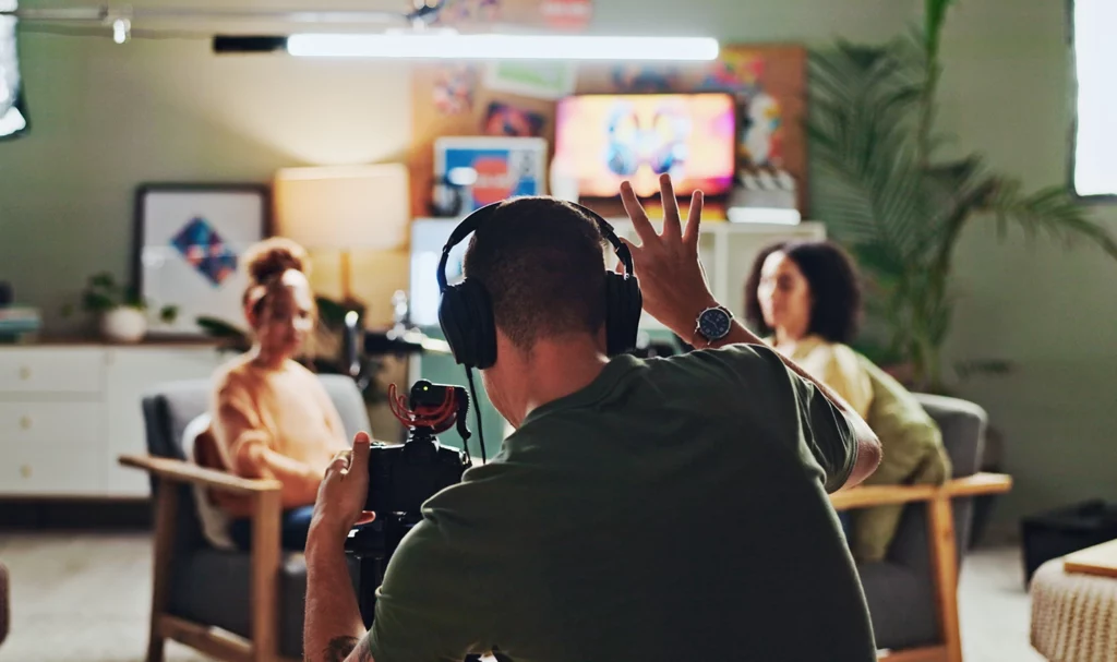 A videographer holds his hands up as he uses his fingers to count down and signal to the two girls sitting across from one another that he is about to start filming their interview.