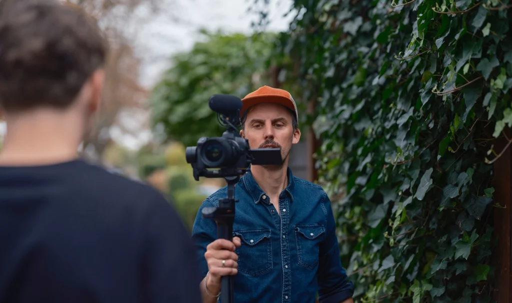 A videographer deep in focus as he films a client speaking in an outdoor space.